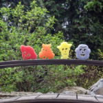 Image Description: All four Toy Story character-shaped Scrub Mommy sponges—red Jessie, orange Bullseye, yellow Woody, and purple Buzz Lightyear—lined up in a row along the dark railing of a wooden bridge, set against a lush green outdoor background of trees and foliage. © Disney/Pixar