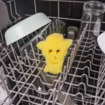 Image Description: A yellow Woody-shaped Scrub Mommy sponge placed upright on the wire rack of an open dishwasher, surrounded by dishes and glassware. © Disney/Pixar