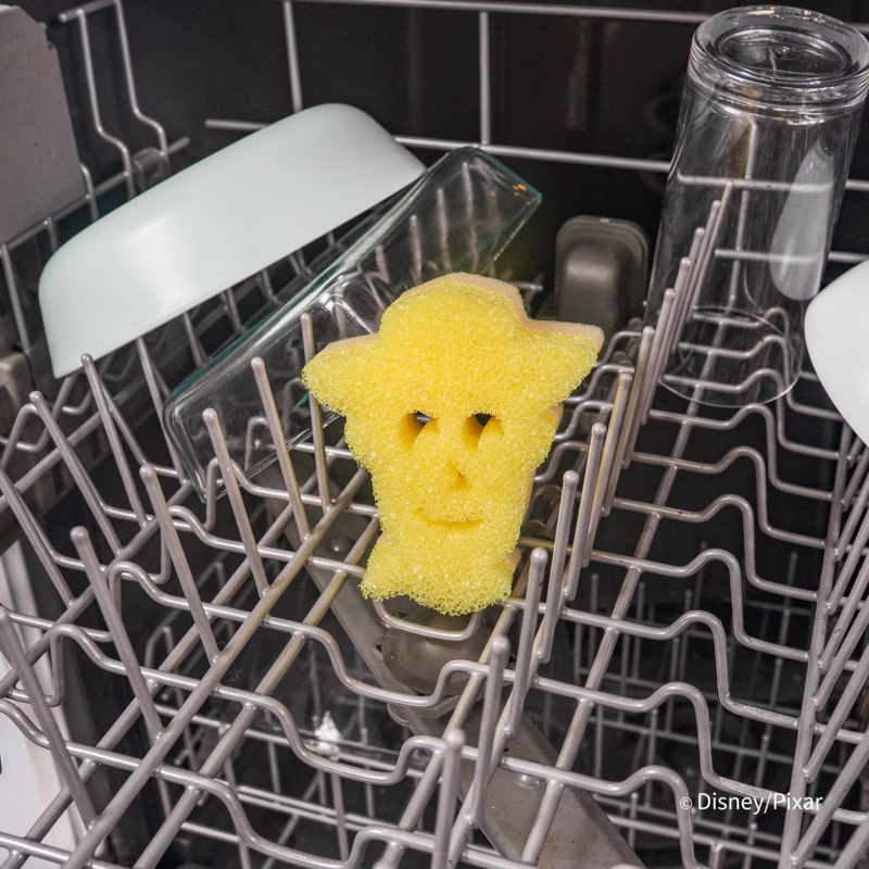Image Description: A yellow Woody-shaped Scrub Mommy sponge placed upright on the wire rack of an open dishwasher, surrounded by dishes and glassware. © Disney/Pixar