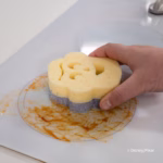 Image Description: A hand holding a purple and yellow Buzz Lightyear-shaped Scrub Mommy sponge, using the textured scrubbing side to clean a messy, stained white stovetop surface. © Disney/Pixar