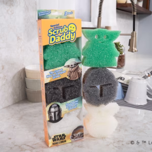 Scrub Daddy Star Wars: The Mandalorian and Grogu Sponges (3-Pack) displayed on a kitchen counter in front of a sink setup. On the left is the special edition retail box featuring Mandalorian branding. Stacked vertically to the right are the three character-shaped sponges: a green Grogu sponge at the top, a dark grey Mandalorian helmet sponge in the middle, and a white Grogu in the hover pram sponge at the bottom. The copyright "© & ™ Lucasfilm Ltd" appears in the bottom right corner.