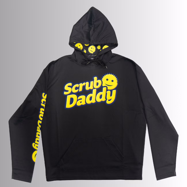 Image Description: A promotional product hero image for a black Scrub Daddy branded hoodie set against a neutral gradient background, featuring the yellow Scrub Daddy logo on the chest, matching yellow branding text down the right sleeve, and a hood interior lined with a yellow smiley face pattern.
