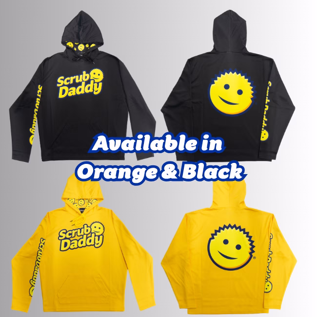 Image Description: A promotional product infographic set against a neutral gradient background, featuring bold blue text in the center that reads "Available in Orange & Black" above and below, showcasing the front and back views of a black Scrub Daddy hoodie on top and a bright orange Scrub Daddy hoodie on the bottom, both featuring the yellow logo on the chest, a large smiley face graphic on the back, and a yellow smiley-patterned hood lining.