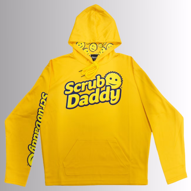 Image Description: A promotional product hero image of a bright orange Scrub Daddy hoodie set against a neutral gradient background, featuring the blue and yellow Scrub Daddy logo on the chest, matching yellow branding down the sleeve, and a hood interior lined with a yellow smiley-face pattern.