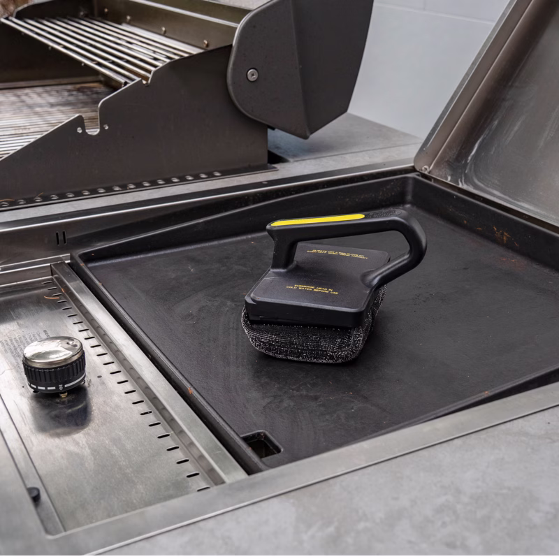 Image Description: Scrub Daddy BBQ Daddy Bristle-Free Griddle Scrubber resting on a black flat-top outdoor griddle next to a grill setup, demonstrating the tool in its natural outdoor cooking environment.
