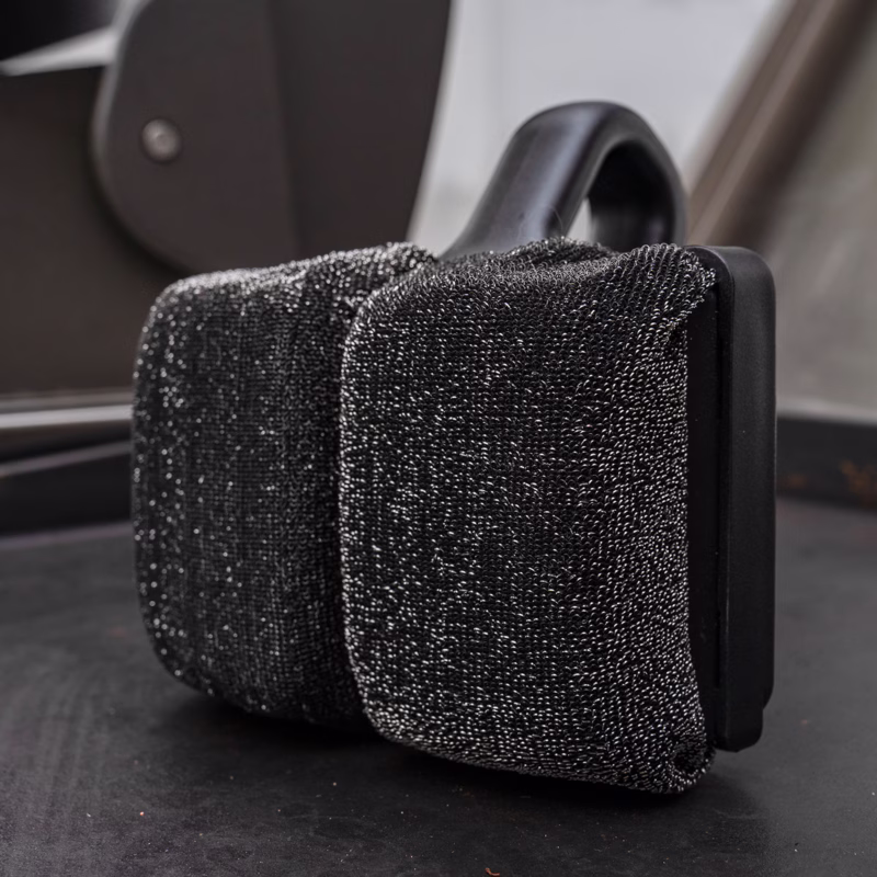 Image Description: Scrub Daddy BBQ Daddy Bristle-Free Griddle Scrubber shown in a close-up shot resting directly on a seasoned black griddle surface, highlighting the textured ArmorTec mesh pads.