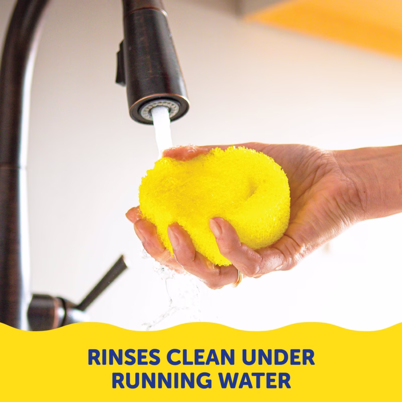 A person's hands holding a yellow Original Scrub Daddy sponge under running water from a kitchen faucet. Text at the bottom reads, "RINSES CLEAN UNDER RUNNING WATER".