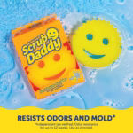 A yellow Original Scrub Daddy sponge and its orange packaging floating in bubbly, soapy blue water. A yellow banner at the bottom contains dark blue text that reads, "RESISTS ODORS AND MOLD*" with smaller text below stating, "*Independent lab verified. Odor resistance for up to 12 weeks. Use as directed."