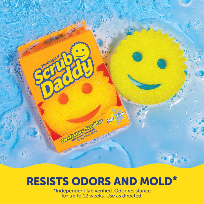 A yellow Original Scrub Daddy sponge and its orange packaging floating in bubbly, soapy blue water. A yellow banner at the bottom contains dark blue text that reads, "RESISTS ODORS AND MOLD*" with smaller text below stating, "*Independent lab verified. Odor resistance for up to 12 weeks. Use as directed."
