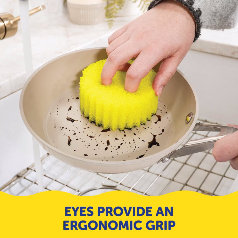 Image Description: A person's hand using the two eye holes of a yellow Scrub Daddy sponge to securely grip it while scrubbing a dirty beige pan in a sink. A yellow banner at the bottom contains dark blue text that reads, "EYES PROVIDE AN ERGONOMIC GRIP".