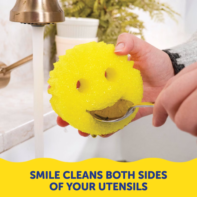 A person holding a yellow Scrub Daddy sponge under a running faucet, using the sponge's smile-shaped mouth to clean both sides of a metal spoon simultaneously. A yellow banner at the bottom contains dark blue text that reads, "SMILE CLEANS BOTH SIDES OF YOUR UTENSILS".