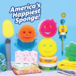 Image Description: A vibrant display of various Scrub Daddy cleaning products arranged on a white tiled surface against a light blue background. The collection includes a Dish Daddy wand, an orange sponge in a clear caddy, pink and yellow sponges stacked on top of a tub of PowerPaste, a wand accessory, a green flower-shaped sponge, and microfiber cloths. White stylized text at the top left reads, "America's Happiest Sponge!".