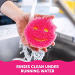 Image Description: A person's hands holding a dual-sided Scrub Mommy sponge, pink side up, under a stream of water from a kitchen faucet. A pink banner at the bottom contains white text reading, "RINSES CLEAN UNDER RUNNING WATER".
