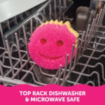 A pink and yellow dual-sided Scrub Mommy sponge resting on the grey tines of a dishwasher rack. A pink banner at the bottom contains white text reading, "TOP RACK DISHWASHER & MICROWAVE SAFE".