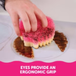 Image Description: A person's hand using the two eye holes of a Scrub Mommy sponge to grip it while scrubbing burnt-on grime off a white stovetop using the pink FlexTexture side. A pink banner at the bottom reads, "EYES PROVIDE AN ERGONOMIC GRIP".
