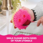Image Description: A person holding a Scrub Mommy sponge under a running faucet, using the sponge's smile-shaped mouth to clean both sides of a metal spoon at the same time. A pink banner at the bottom reads, "SMILE CLEANS BOTH SIDES OF YOUR UTENSILS".