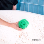 A person uses a green Oogie Boogie Scrub Daddy sponge to clean a white surface splattered with reddish brown spots against a blue tiled wall. The Disney copyright text appears in the bottom right.