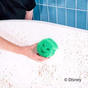 Image Description: A person uses a green Oogie Boogie Scrub Daddy sponge to clean a white surface splattered with reddish brown spots against a blue tiled wall. The Disney copyright text appears in the bottom right.