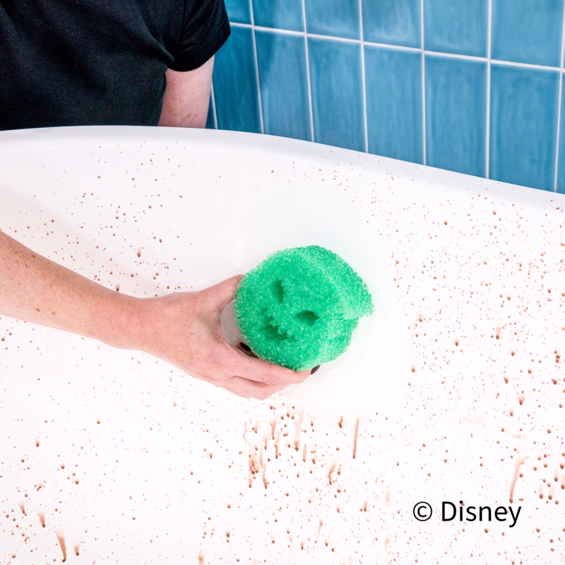 A person uses a green Oogie Boogie Scrub Daddy sponge to clean a white surface splattered with reddish brown spots against a blue tiled wall. The Disney copyright text appears in the bottom right.