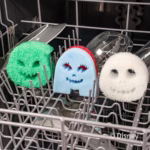 Image Description: Three character-themed Scrub Daddy and Scrub Mommy sponges (green Oogie Boogie, blue Sally, and white Jack Skellington) resting on the grey wire tines of an open dishwasher rack. The Disney copyright text appears in the bottom right.