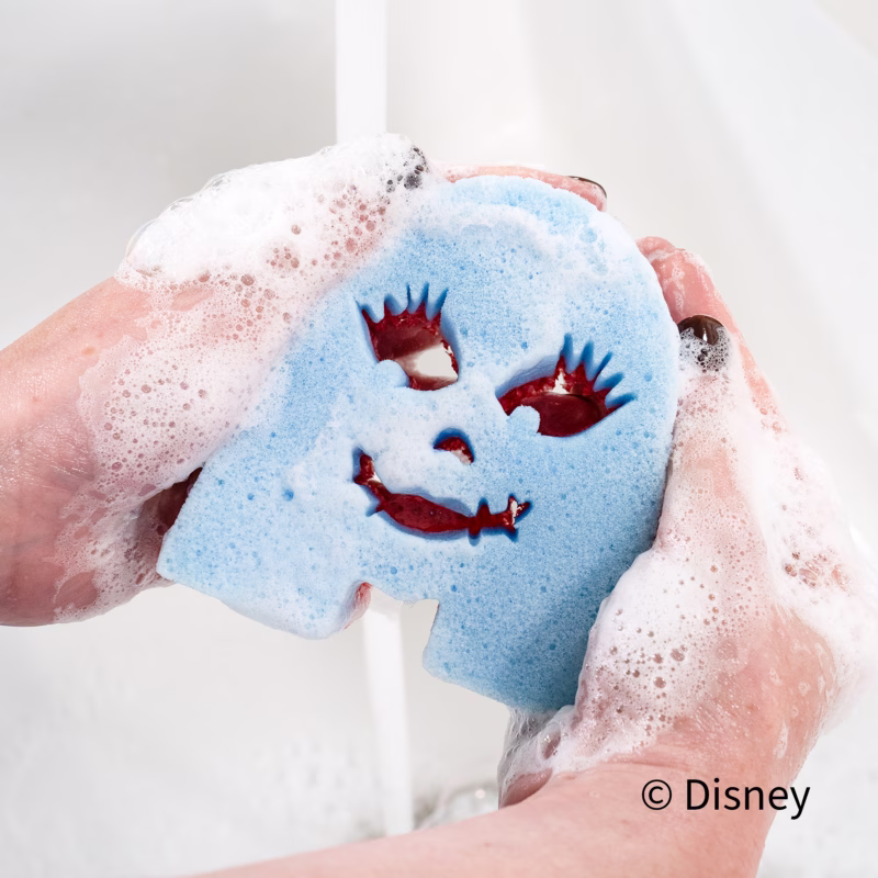 person's soapy hands hold a blue, dual-sided Sally Scrub Mommy sponge under running water in a sink, showing suds and the character's facial features. The Disney copyright text appears in the bottom right.