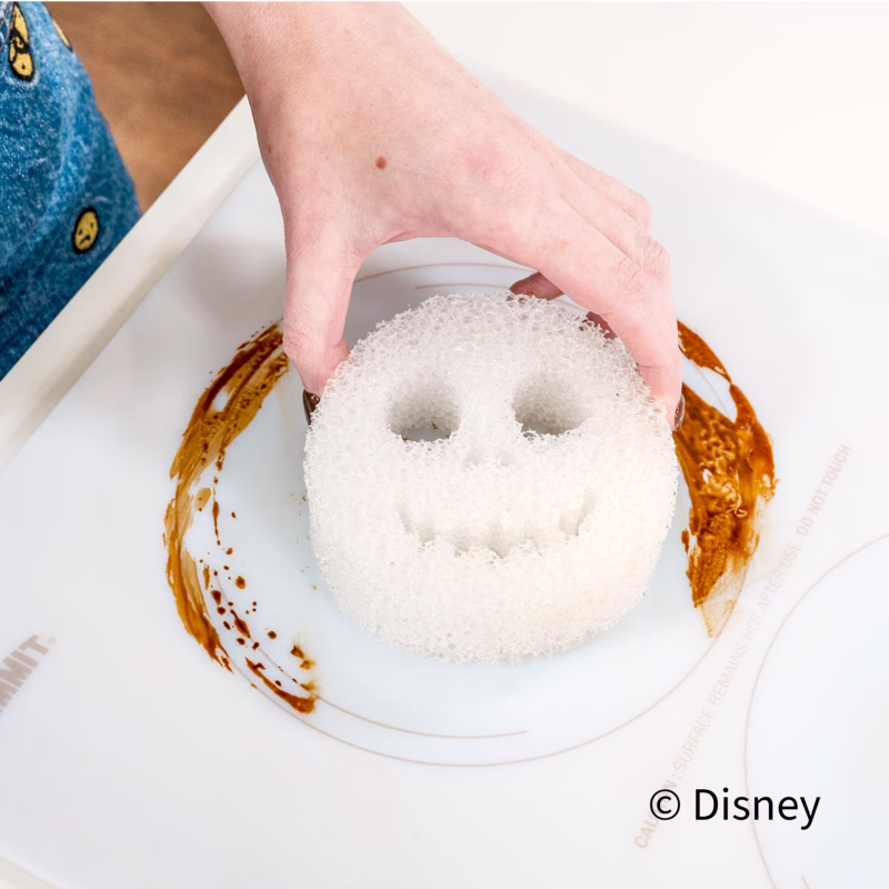 A person uses a white Jack Skellington Scrub Daddy sponge to clean a dirty white stovetop ring, leaving a clean track through the burnt residue. The Disney copyright text appears in the bottom right.