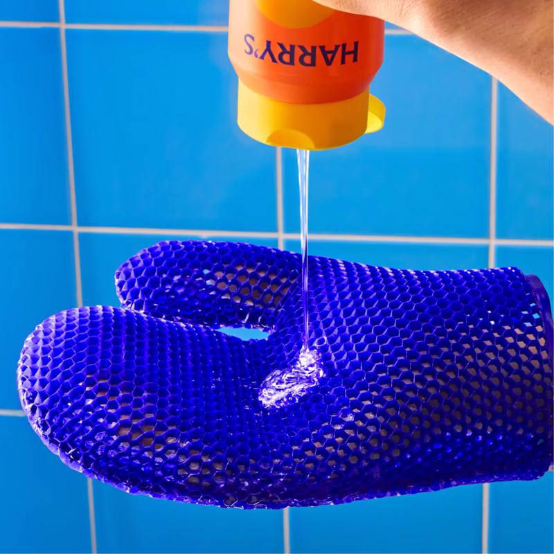 Image Description: An action product photo showing body wash being poured from the bottle onto the textured surface of the blue exfoliating mitt against a blue tiled background.