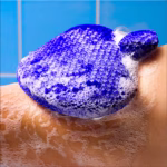Image Description: A close-up lifestyle action photo of the soapy blue exfoliating mitt being used on skin during a shower against a blue tiled background.