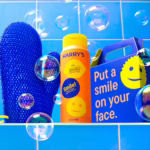 Image Description: A lifestyle product photo of the Harry's x Scrub Daddy full kit set against a blue tiled wall background filled with floating soap bubbles, showing the blue exfoliating mitt, the orange Lemon & Mint body wash bottle, and the blue gift box.