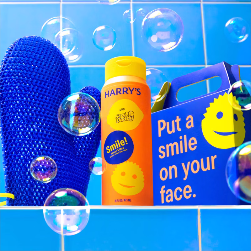 Image Description: A lifestyle product photo of the Harry's x Scrub Daddy full kit set against a blue tiled wall background filled with floating soap bubbles, showing the blue exfoliating mitt, the orange Lemon & Mint body wash bottle, and the blue gift box.