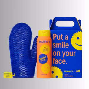 Image Description: A promotional product hero image of the Harry's x Scrub Daddy collaboration set against a neutral gradient background, featuring a blue exfoliating mitt, a 16 fl oz bottle of Lemon & Mint body wash, and a branded blue gift box with yellow smiley graphics.