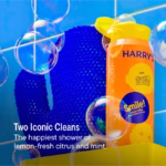 Image Description: A product feature infographic set against a blue tiled background with floating soap bubbles, featuring the Harry's x Scrub Daddy body wash and blue exfoliating mitt, highlighting "Two Iconic Cleans" and a lemon-fresh citrus and mint scent.