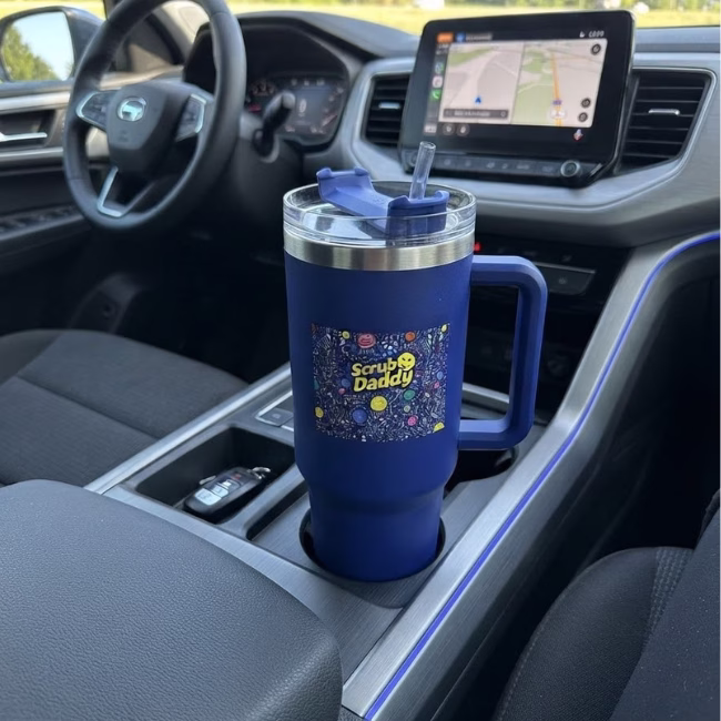 Image Description: A lifestyle product photo showing the royal blue Scrub Daddy travel tumbler sitting securely in the center console cup holder of a vehicle, with the dashboard, steering wheel, and a GPS navigation screen visible in the background.