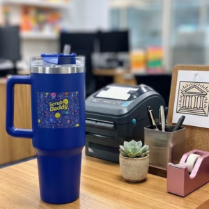 Image Description: A lifestyle product photo of the royal blue Scrub Daddy travel tumbler placed upright on an office desk next to a small succulent plant, pen holder, tape dispenser, and a receipt printer in a bright workspace.