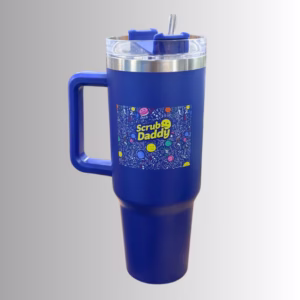 Image Description: A promotional product hero image of a royal blue insulated travel tumbler set against a neutral gradient background, featuring a sturdy side handle, a clear lid with a blue straw and sliding closure, and a vibrant multi-colored doodle graphic square with the "Scrub Daddy" logo on the front.