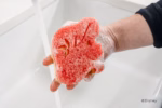 Image Description: A lifestyle action photo showing a hand squeezing soapy suds from the red Ariel Disney Princess Scrub Mommy sponge over a kitchen sink.