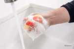 Image Description: A lifestyle action photo of a hand holding the red Ariel Disney Princess Scrub Mommy sponge under a running kitchen faucet.