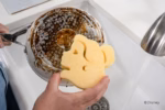 Image Description: A lifestyle action photo of a hand scrubbing the bottom of a soiled pot with the yellow Belle Disney Princess Scrub Mommy sponge in a kitchen sink.