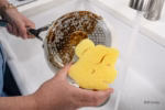 Image Description: A lifestyle action photo showing a hand holding the yellow Belle Disney Princess Scrub Mommy sponge next to a dirty pot in a kitchen sink.