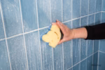Image Description: A lifestyle action photo showing a hand wiping down soapy blue bathroom tiles with the yellow side of the Cinderella Disney Princess Scrub Mommy sponge.