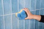 Image Description: A lifestyle action photo showing a hand using the blue Cinderella Disney Princess Scrub Mommy sponge to clean soapy blue bathroom tiles.